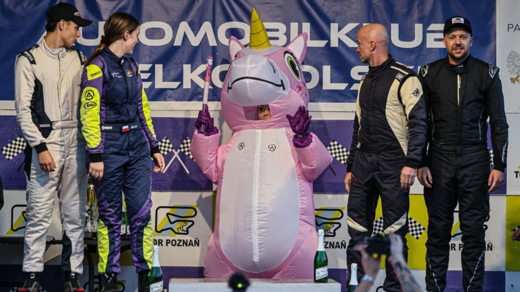 Ceremonia podium R Sport Cup na Torze Poznań — kierowcy w kombinezonach obok postaci w kostiumie jednorożca